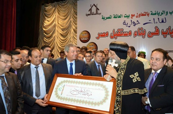 Pope Tawadros presenting Minister of Youth Khalid Abdel Aziz with a commemorative gift, quoting Isaiah 19:25 in Coptic, Arabic, and English.
